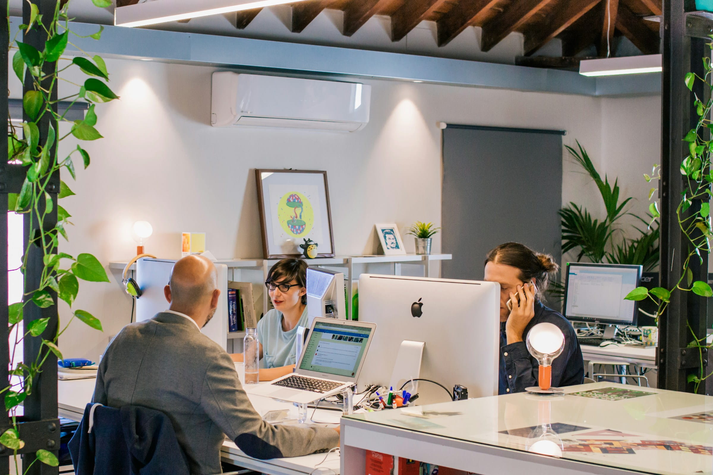 Three people work at their desks in a modern office setting. One person is on the phone, while two others are facing each other, seemingly in discussion.