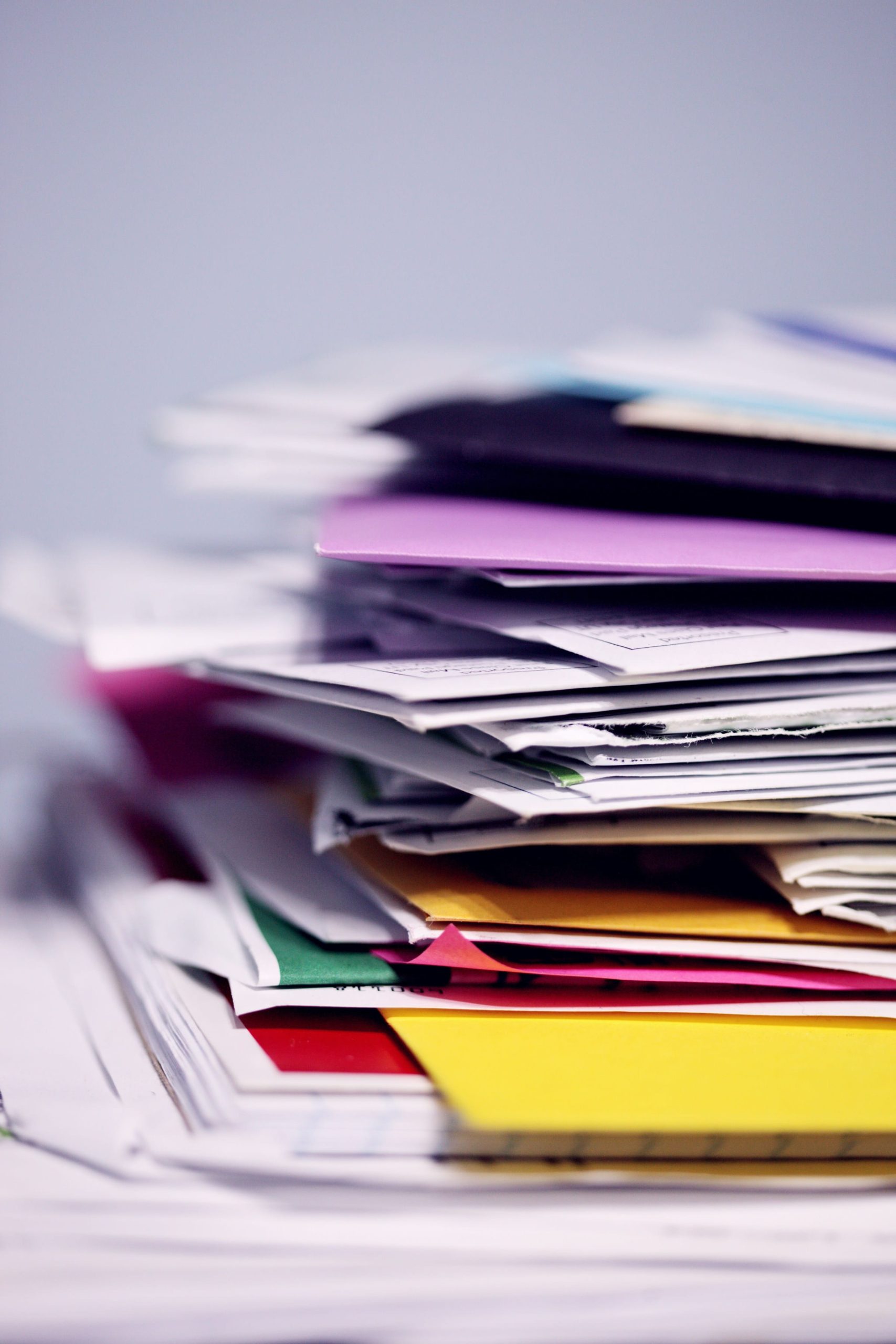 A tall stack of papers and folders of various colors is shown, with the background out of focus. The papers are piled unevenly, creating a sense of disarray.