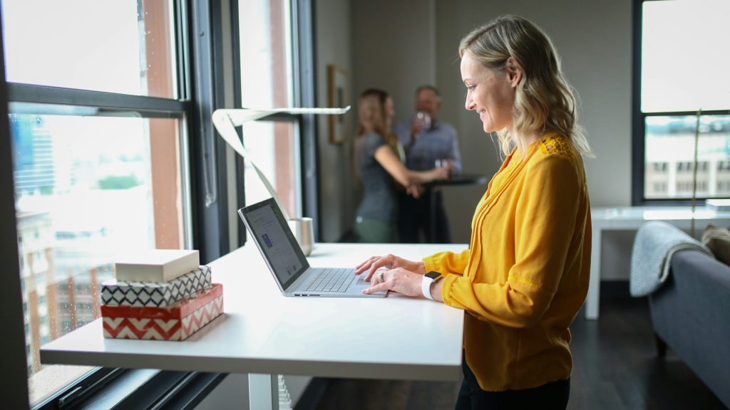 A woman in a yellow shirt smiles as she uses a laptop at a standing desk. In the background, two other people are conversing.