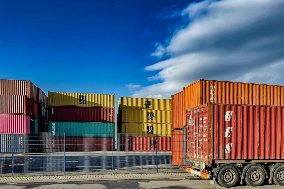 A truck is positioned in front of a stack of colorful shipping containers under a bright blue sky. The containers are various colors including red, yellow, pink, and teal, and are neatly stacked in rows.