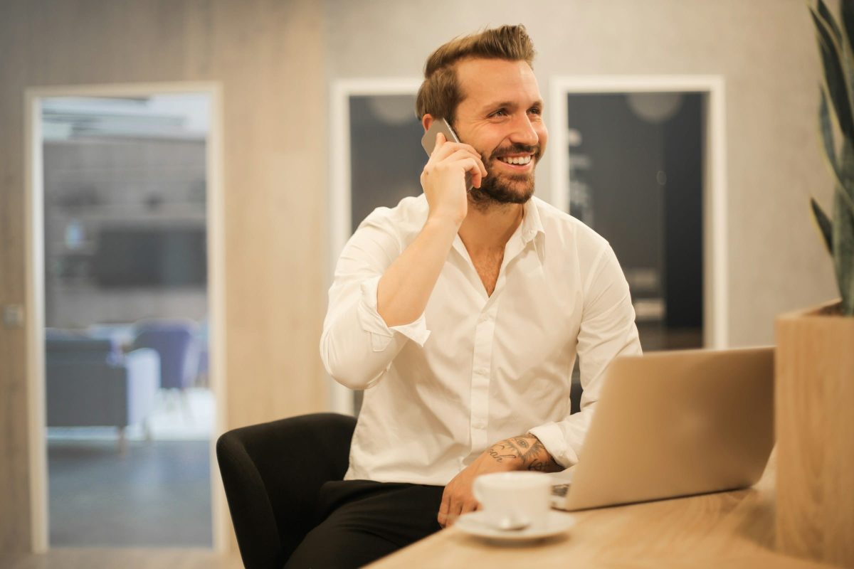 A smiling man in a white shirt is on the phone at a desk with a laptop. He is seated in front of a plant and a cup of coffee.