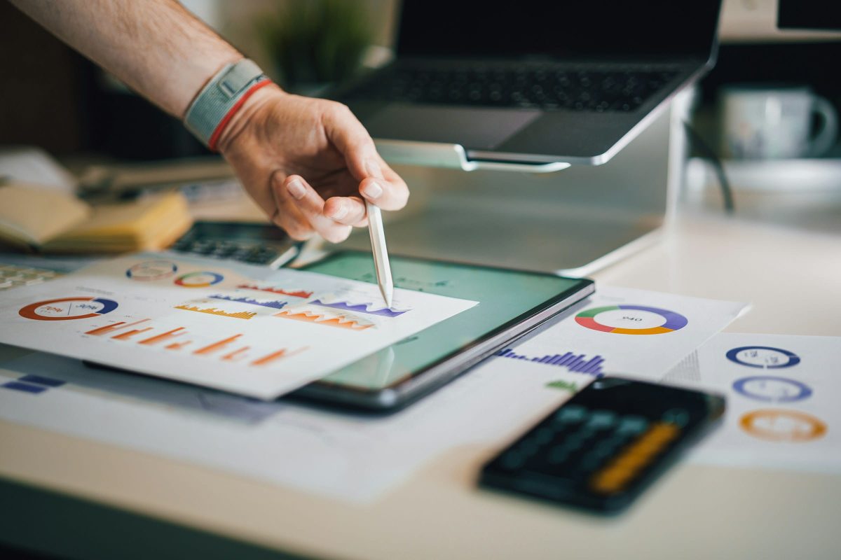 A person's hand holds a stylus over a tablet displaying financial charts. The tablet is on a desk with scattered financial documents and a laptop in the background.