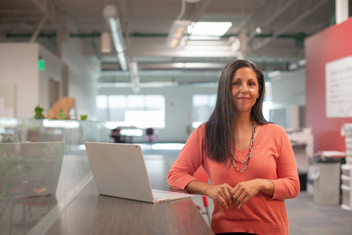 A smiling woman with long dark hair stands in an office with a laptop in front of her. She is wearing an orange sweater and a beaded necklace.
