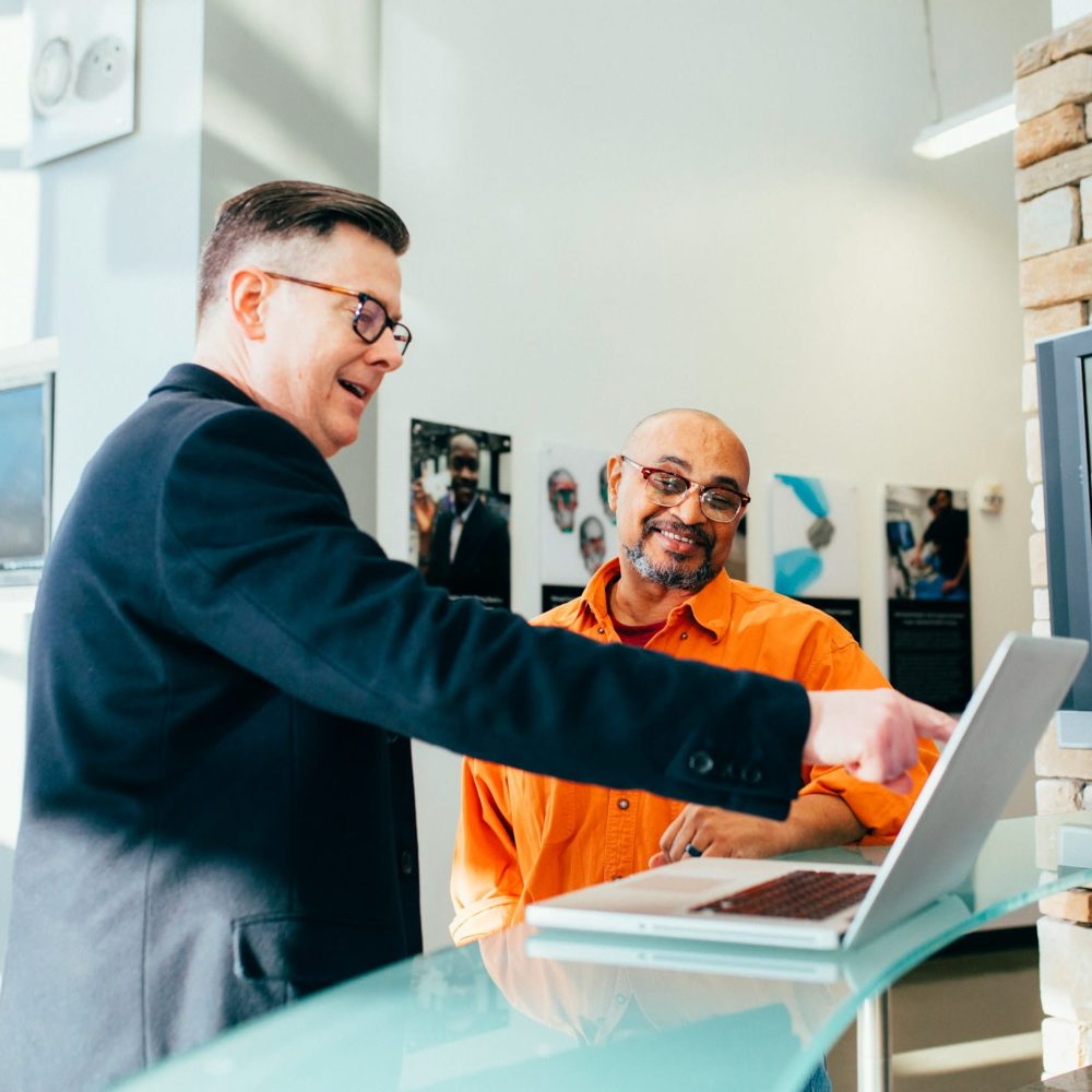 Two men, one in a suit jacket and the other in an orange shirt, are looking at a laptop on a glass table. The man in the suit jacket is pointing at the laptop screen, and both men are smiling.