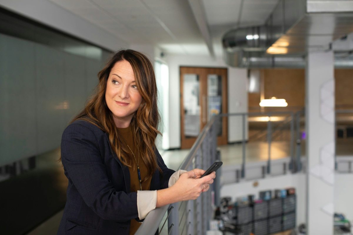 A woman in a blazer and brown shirt leans on a railing while holding a smartphone. The background shows a modern office environment with exposed ductwork and lighting.