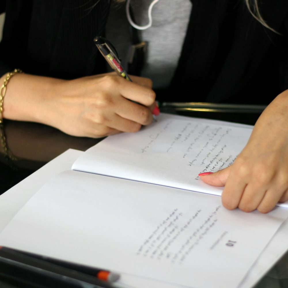 A person's hands are visible as they write on a document with a pen. One hand wears a bracelet and the other a watch.