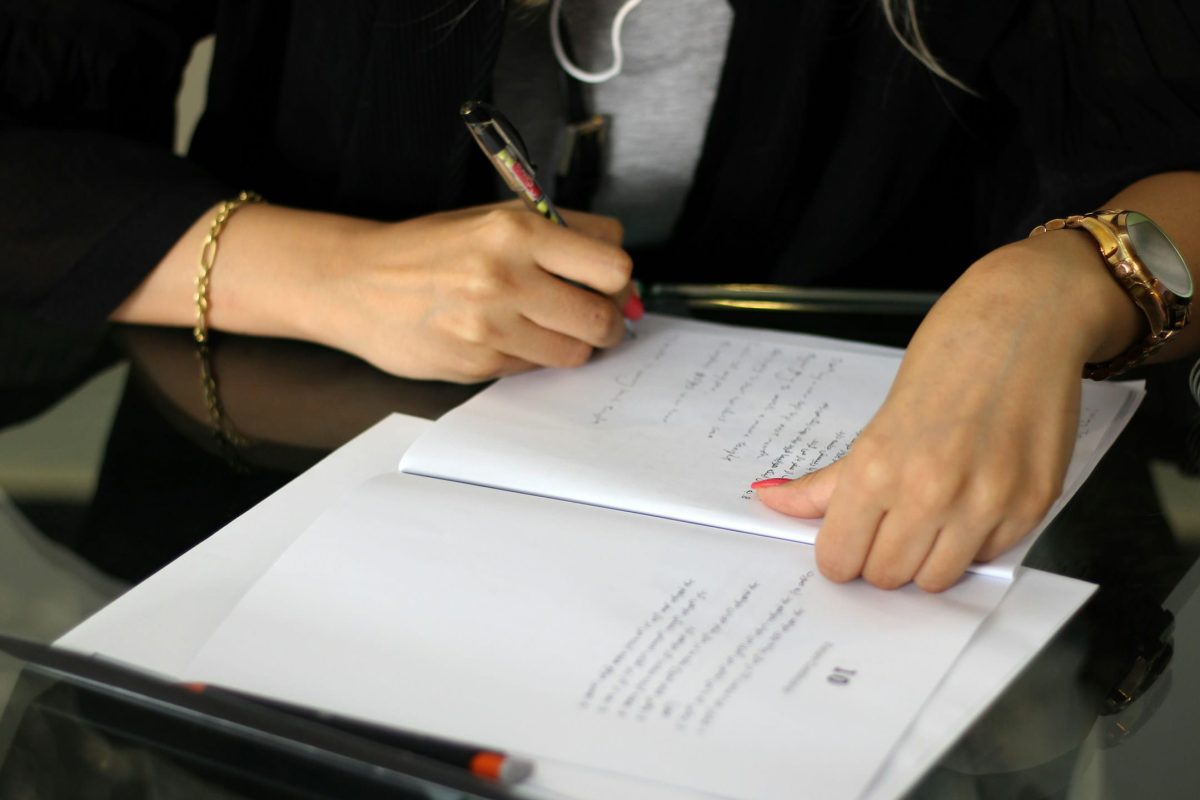 A person's hands are visible as they write on a document with a pen. One hand wears a bracelet and the other a watch.