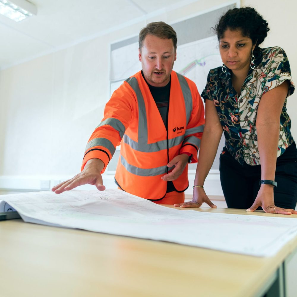 A man in an orange safety vest and a woman in a patterned shirt are looking at blueprints on a table. The man is pointing to the plans.