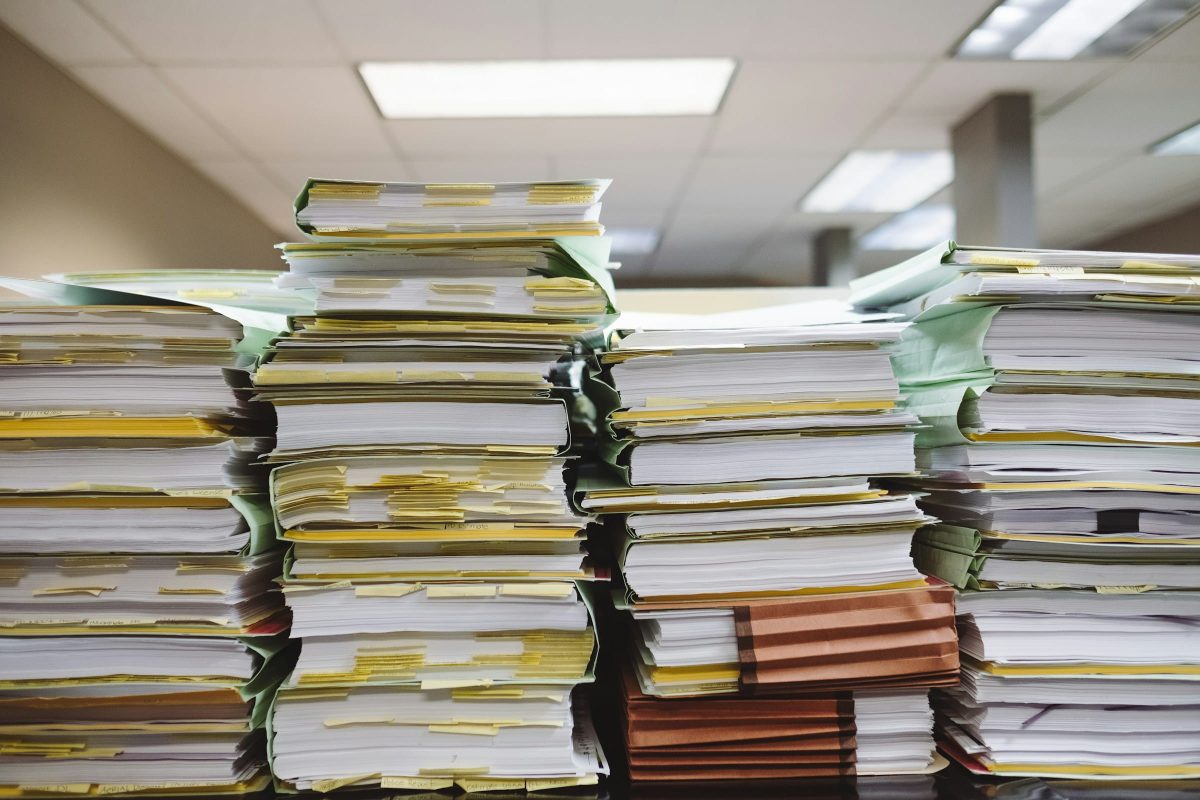 Tall stacks of papers are organized in folders and piled high on a desk. The folders are a mix of green and yellow, with some brown file folders also visible.
